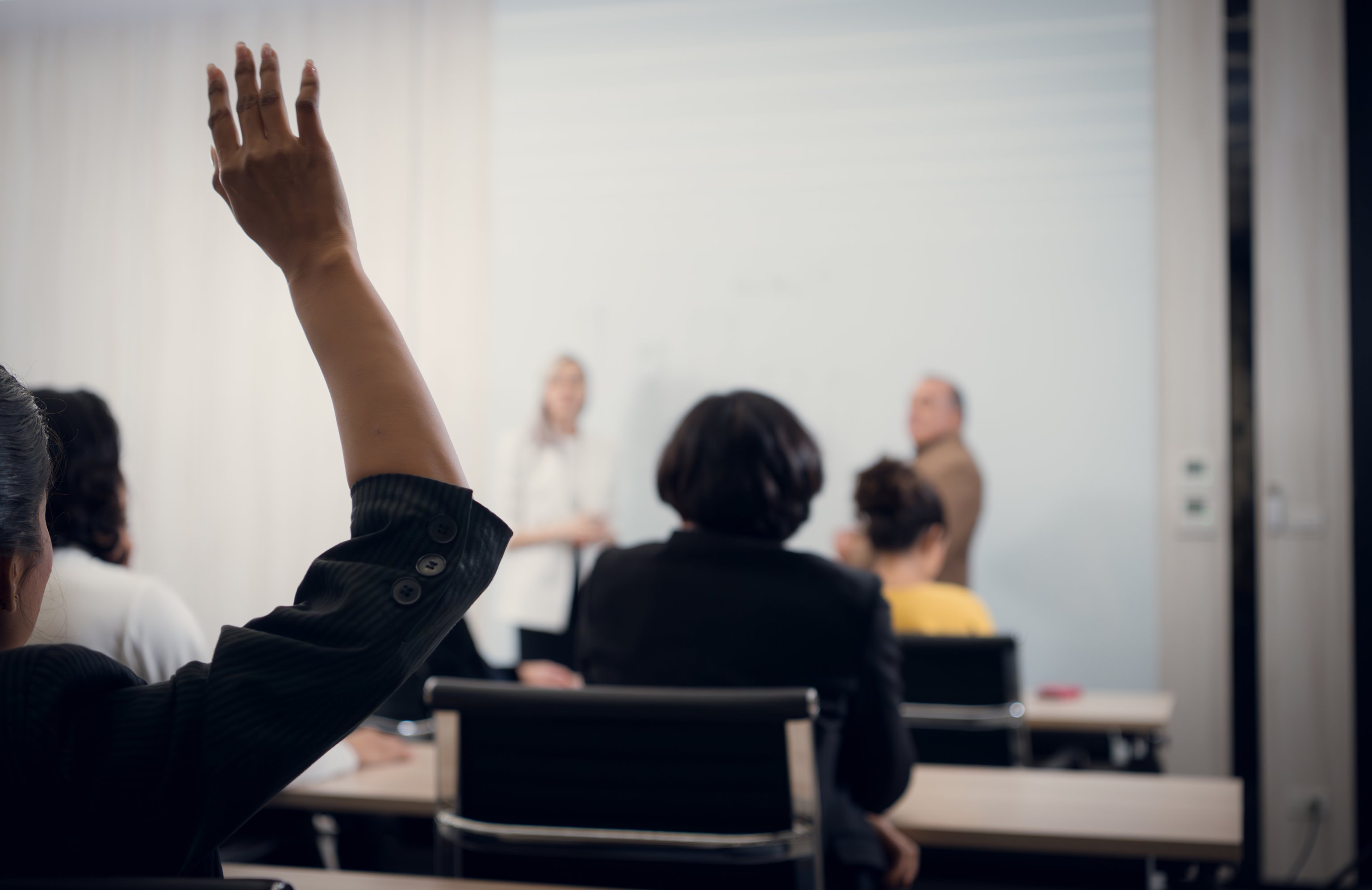 A person raising their hand in a classroom or lecture hall setting with other attendees seated at tables, actively participating in a presentation or discussion.