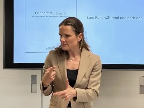A woman standing behind a presentation desk gesturing as she speaks, with a slide projector and presentation screen visible in the background.