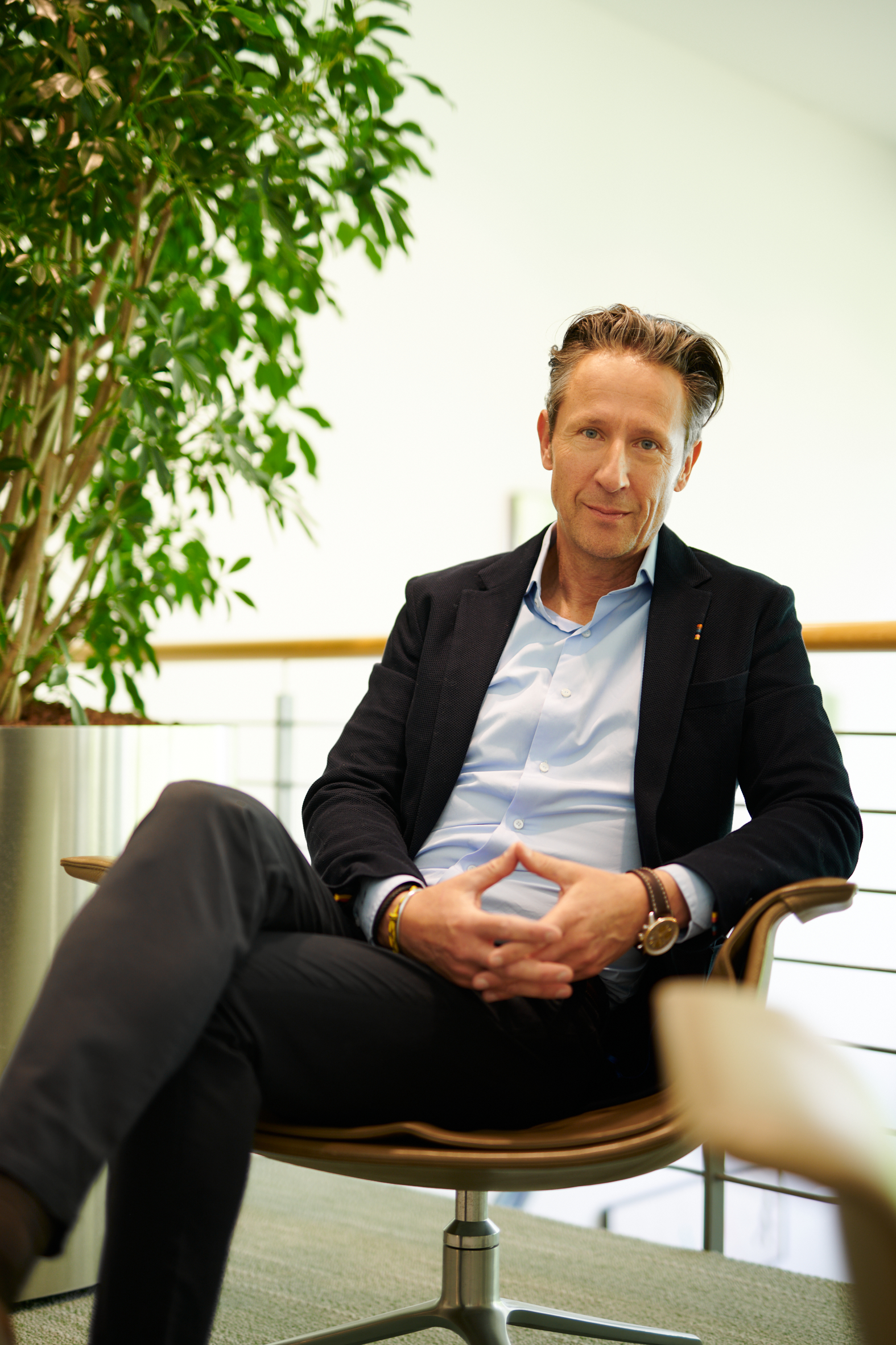 A man in a dark blue suit sitting in a chair with legs crossed, with a contemplative expression and a plant visible to the left of the frame.